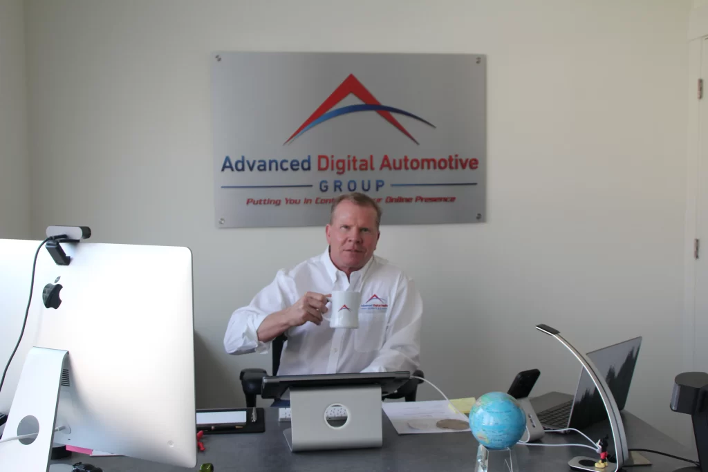 The CEO of Advanced Digital Automotive Group sits at his desk, holding a mug, with the company logo behind.